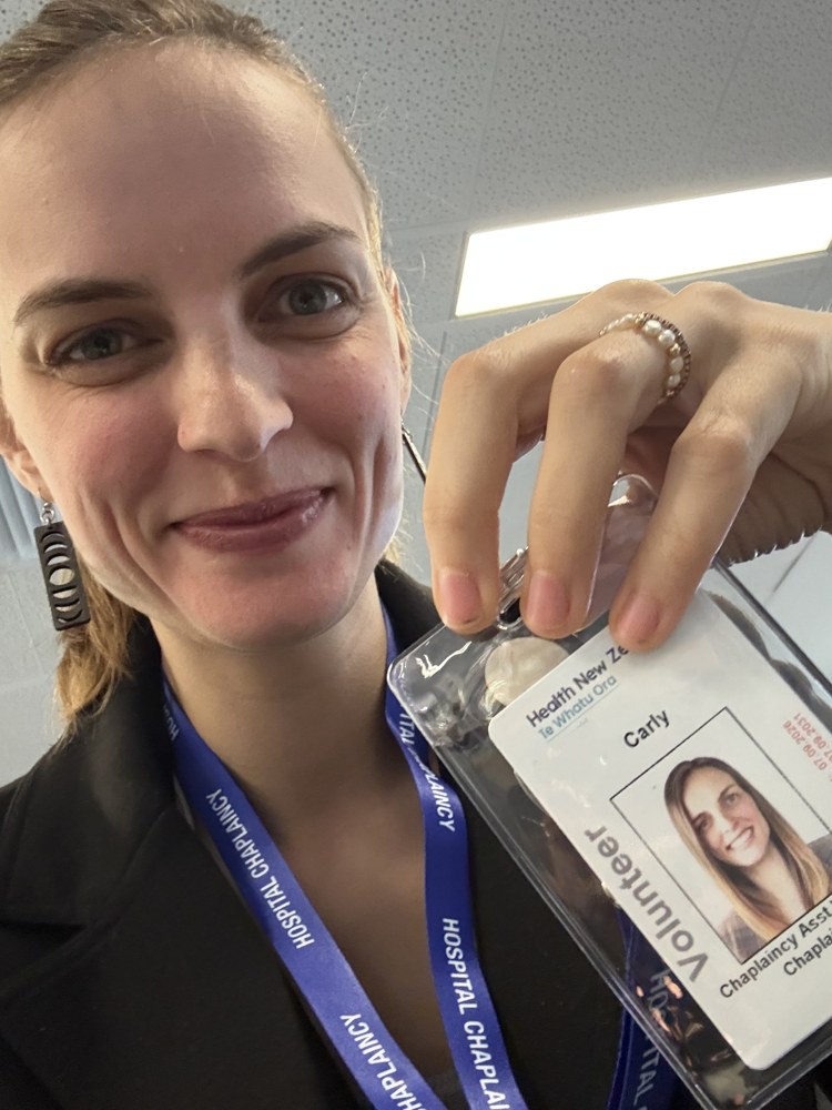 Carly smiling in a blazer, holding up her hospital volunteer badge on a Hospital Chaplaincy lanyard