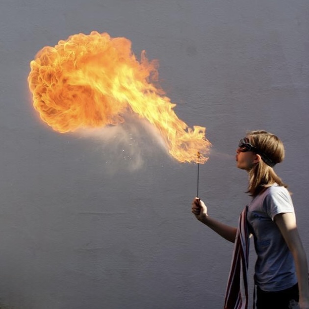 Carly breathing a large ball of fire against a grey wall, wearing goggles.